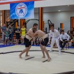 Young athletes engaged in sumo wrestling in a gym with spectators and a referee observing.