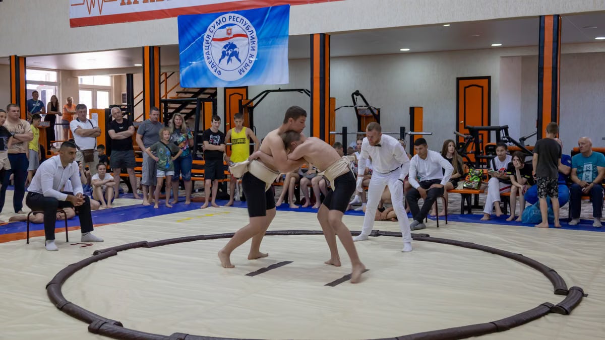 Young athletes engaged in sumo wrestling in a gym with spectators and a referee observing.